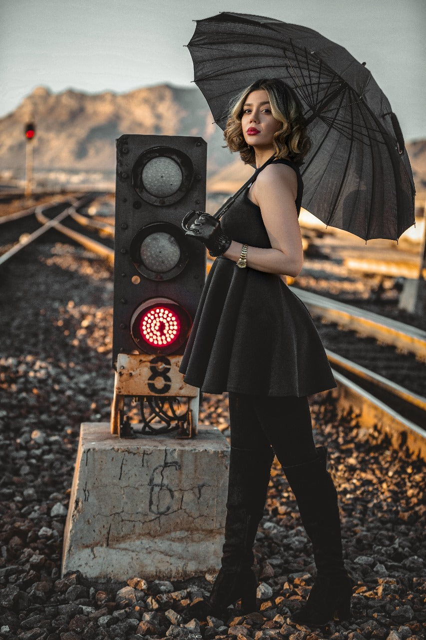 Woman wearing XAYWEAR black dress holding black umbrella standing by railway tracks