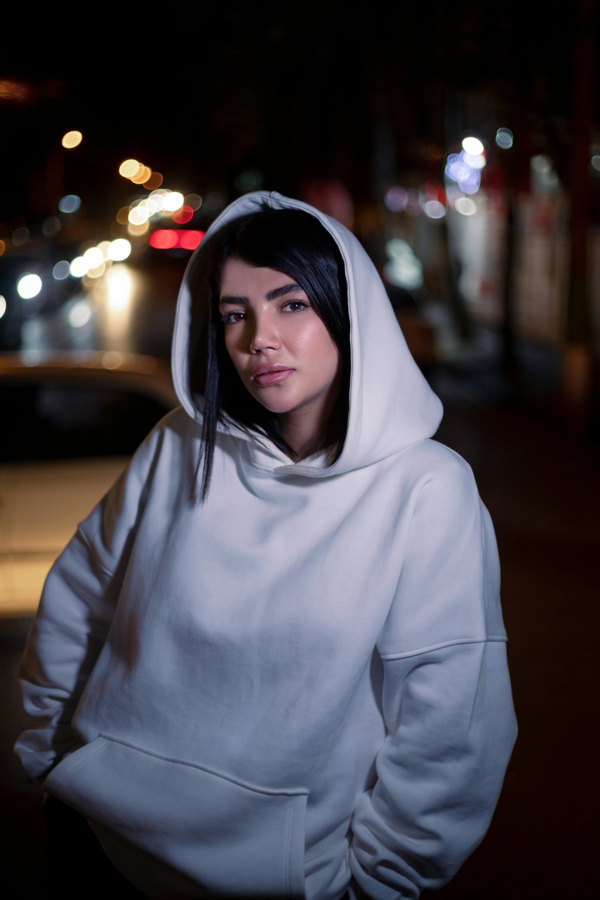 XAYWEAR white hoodie worn by woman standing on city street at night with blurred lights